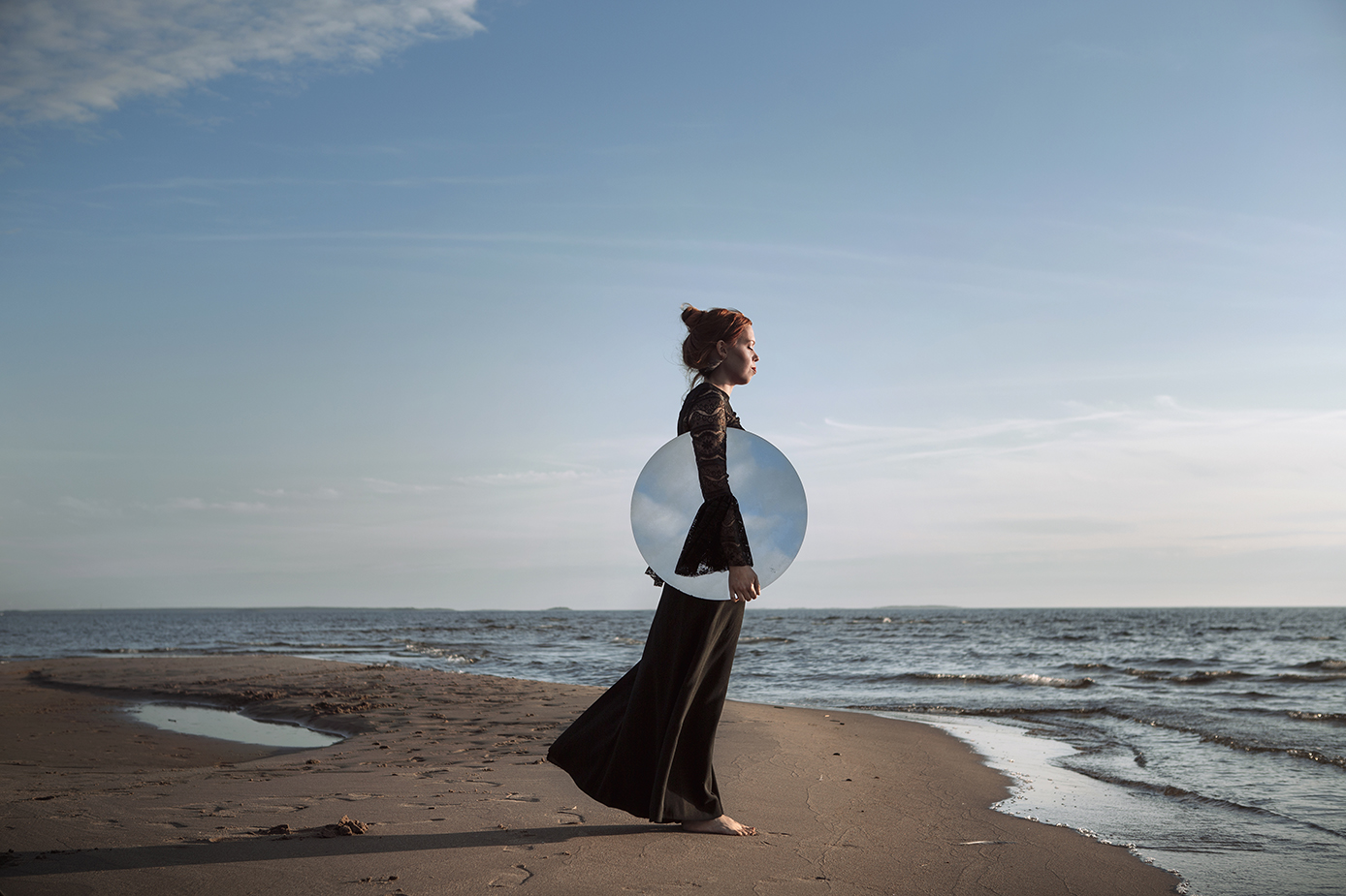 Nainen seisoo aavan meren rannalla silmät kiinni kasvot auringon valoa kohti. Kainalossaan hänellä on pyöreä peili, josta heijastuu sininen taivas ja pilvenhahtuvat.