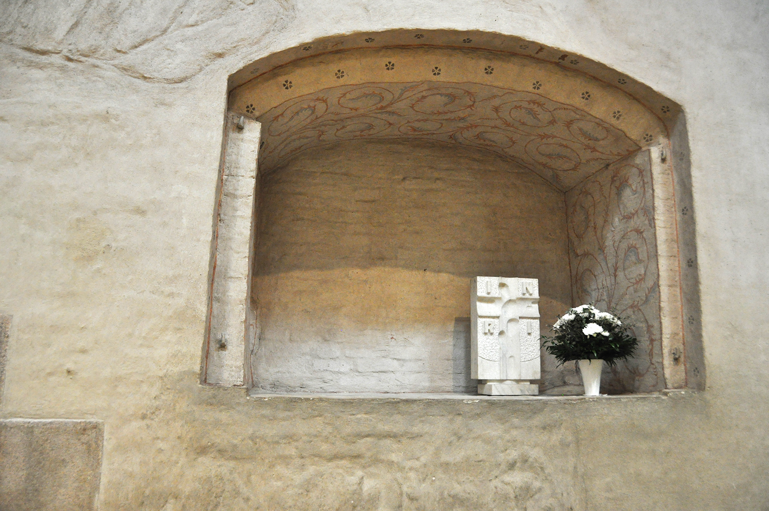 Kaarisyvennys kirkon sisäseinässä, syvennyksessä valkoinen kukkakimppu ja marmorikrusifiksi