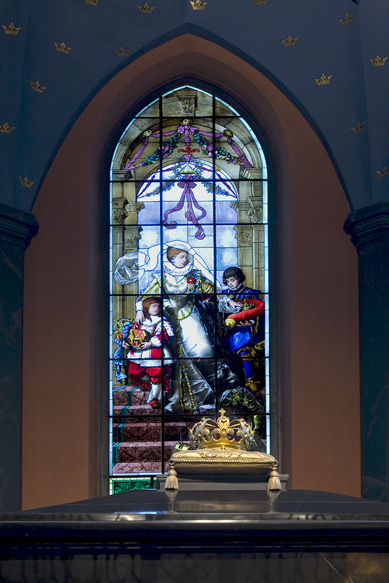 Taustalla lasimaalaus, jossa nainen ja kaksi poikaa, etualalla musta sarkofagi, jonka päällä kruunu